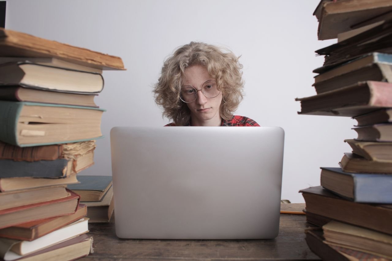 Services Concentrated male teenager in casual shirt and eyeglasses sitting at wooden table with books and using laptop while preparing for exams